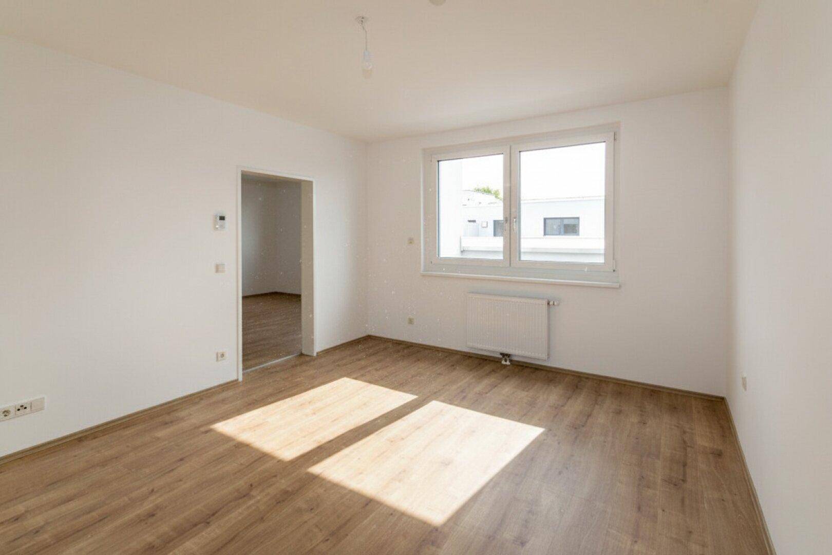 Lichtdurchfluteter Raum mit Laminatboden, großen Fenstern und offenem Zugang zu einem angrenzenden Bereich.