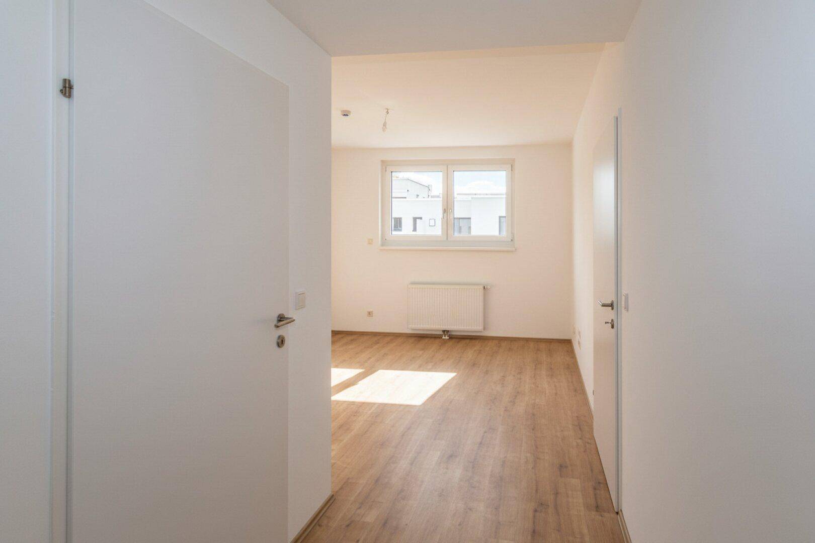 Langer, heller Flur mit Laminatboden, der zu einem weiteren lichtdurchfluteten Zimmer führt.