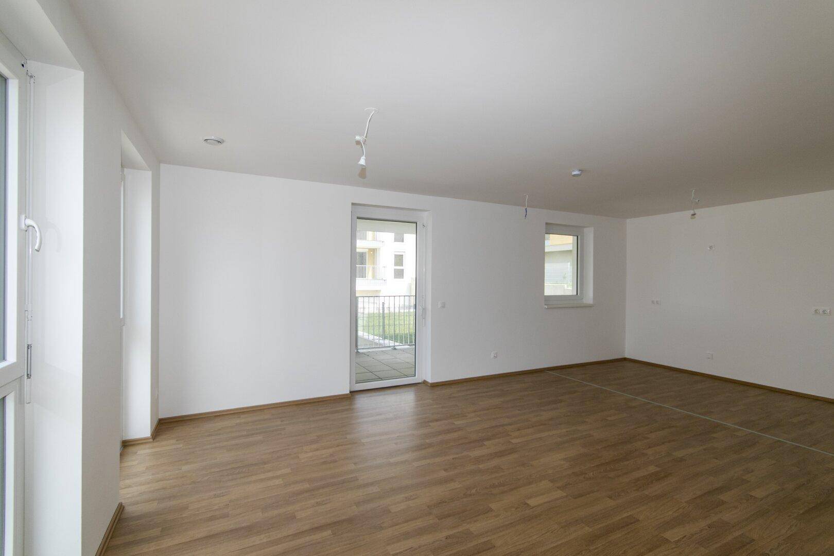 Geräumiges, unmöbliertes Wohnzimmer mit Laminatboden und großen Fenstern für viel Tageslicht.