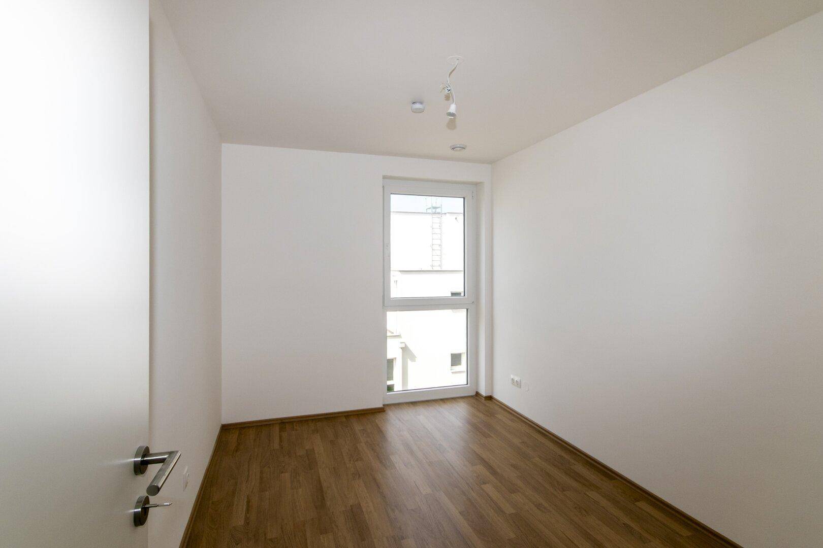 Unmöbliertes Schlafzimmer mit einem großen Fenster, das viel Tageslicht in den Raum lässt.