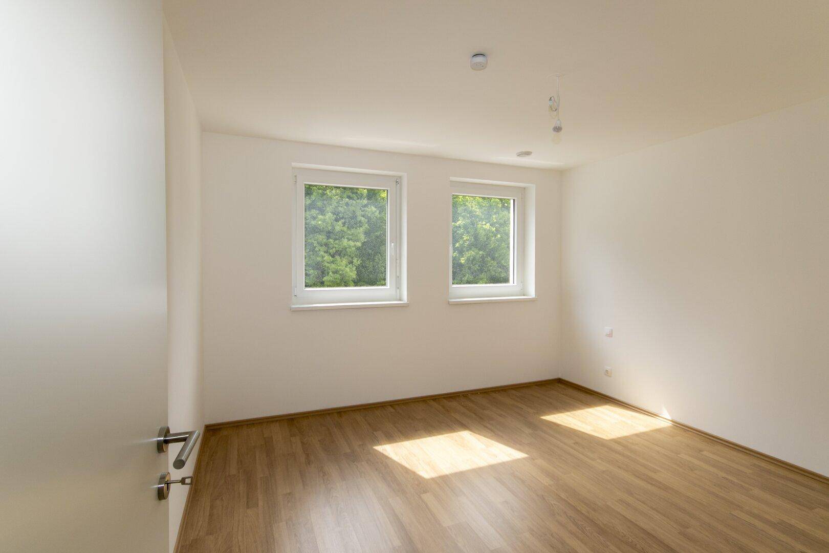 Helles Schlafzimmer mit Laminatboden und zwei Fenstern mit Blick ins Grüne.