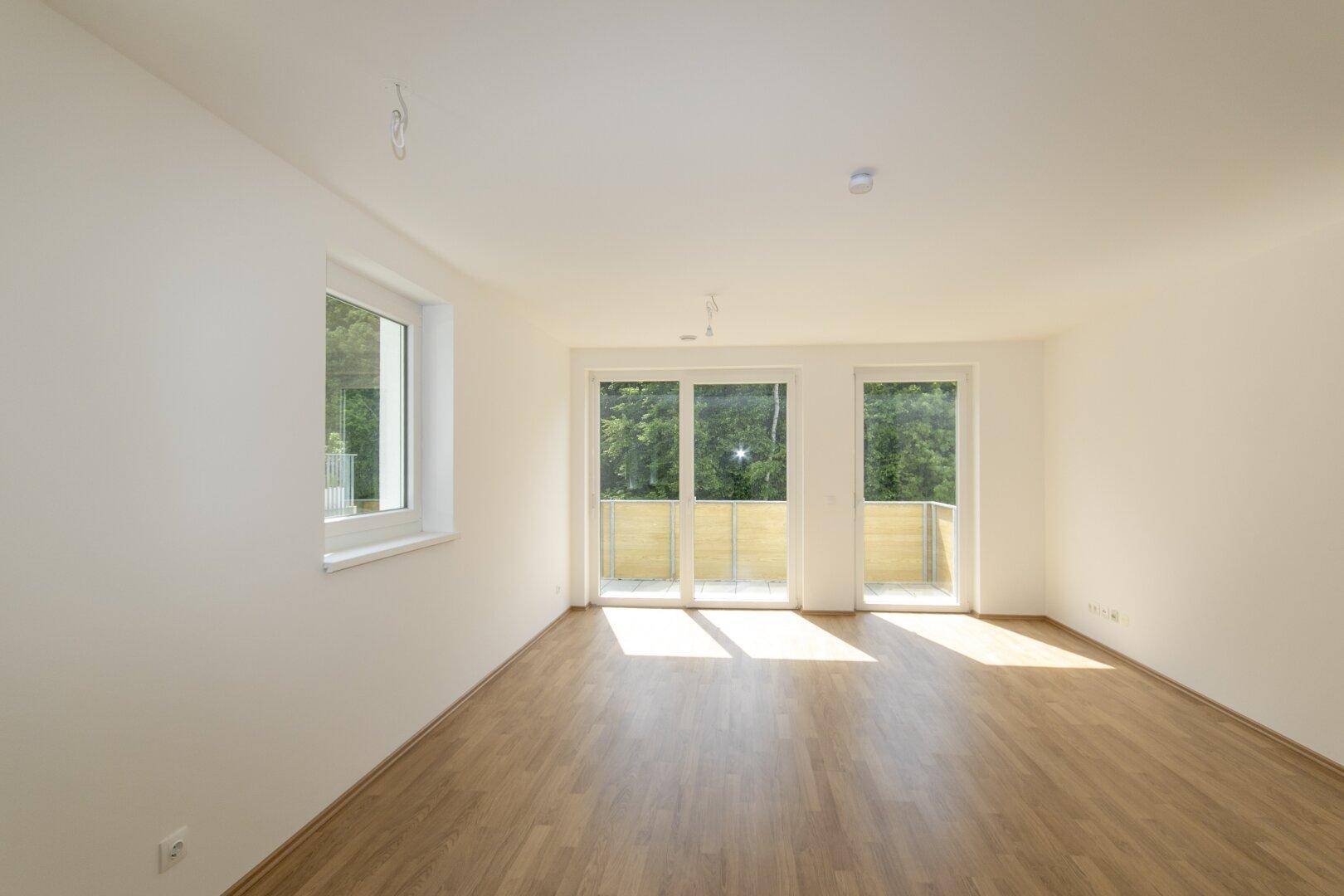 Helles, leeres Zimmer mit Laminatboden und Zugang zu einem Balkon mit Waldblick.