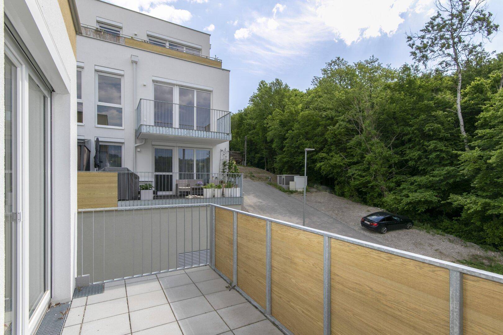 Balkon mit Blick auf das moderne Gebäude und die bewaldete Umgebung unter blauem Himmel.