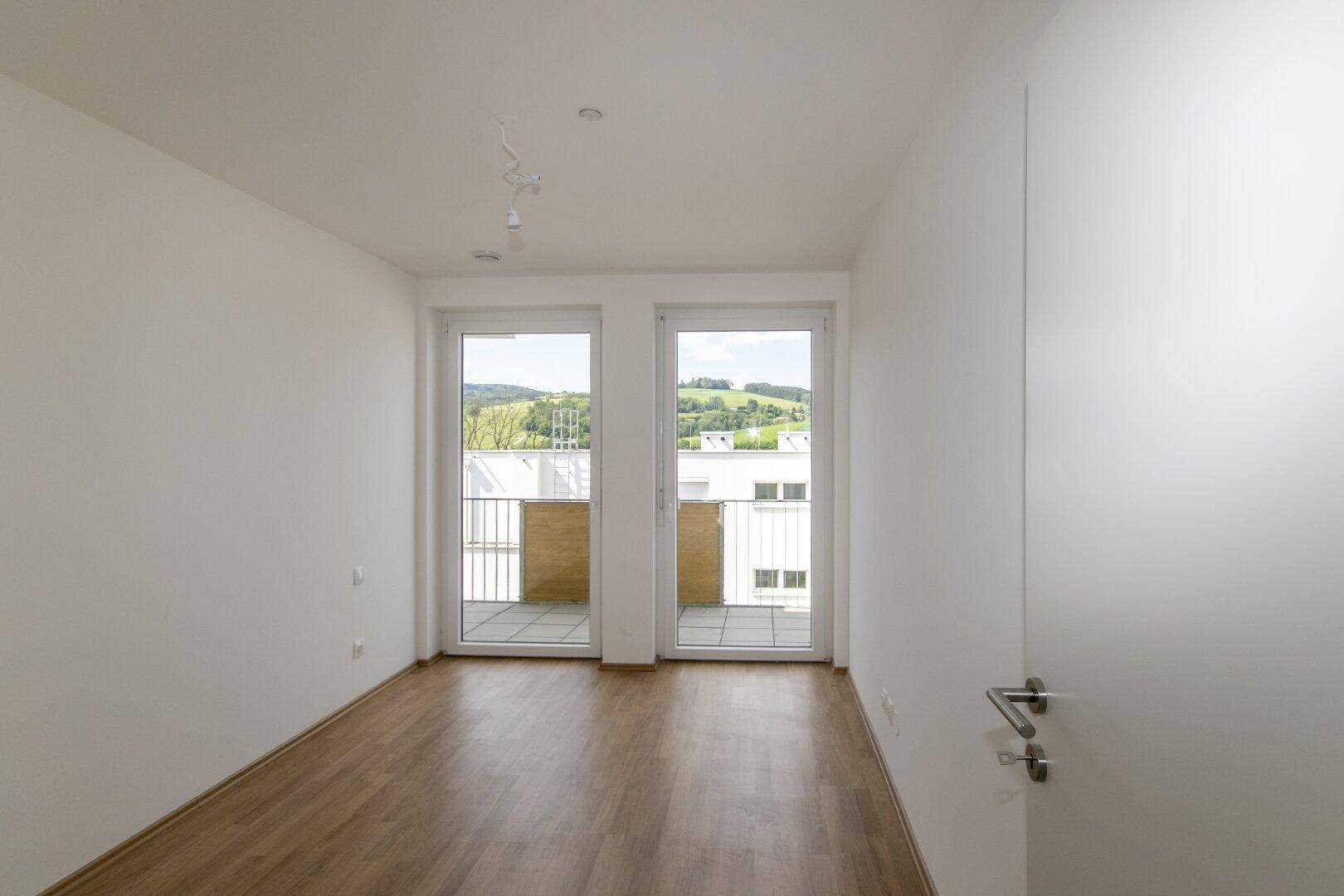 Helles Schlafzimmer mit Laminatboden und zwei Fenstertüren zum Balkon mit grüner Aussicht.
