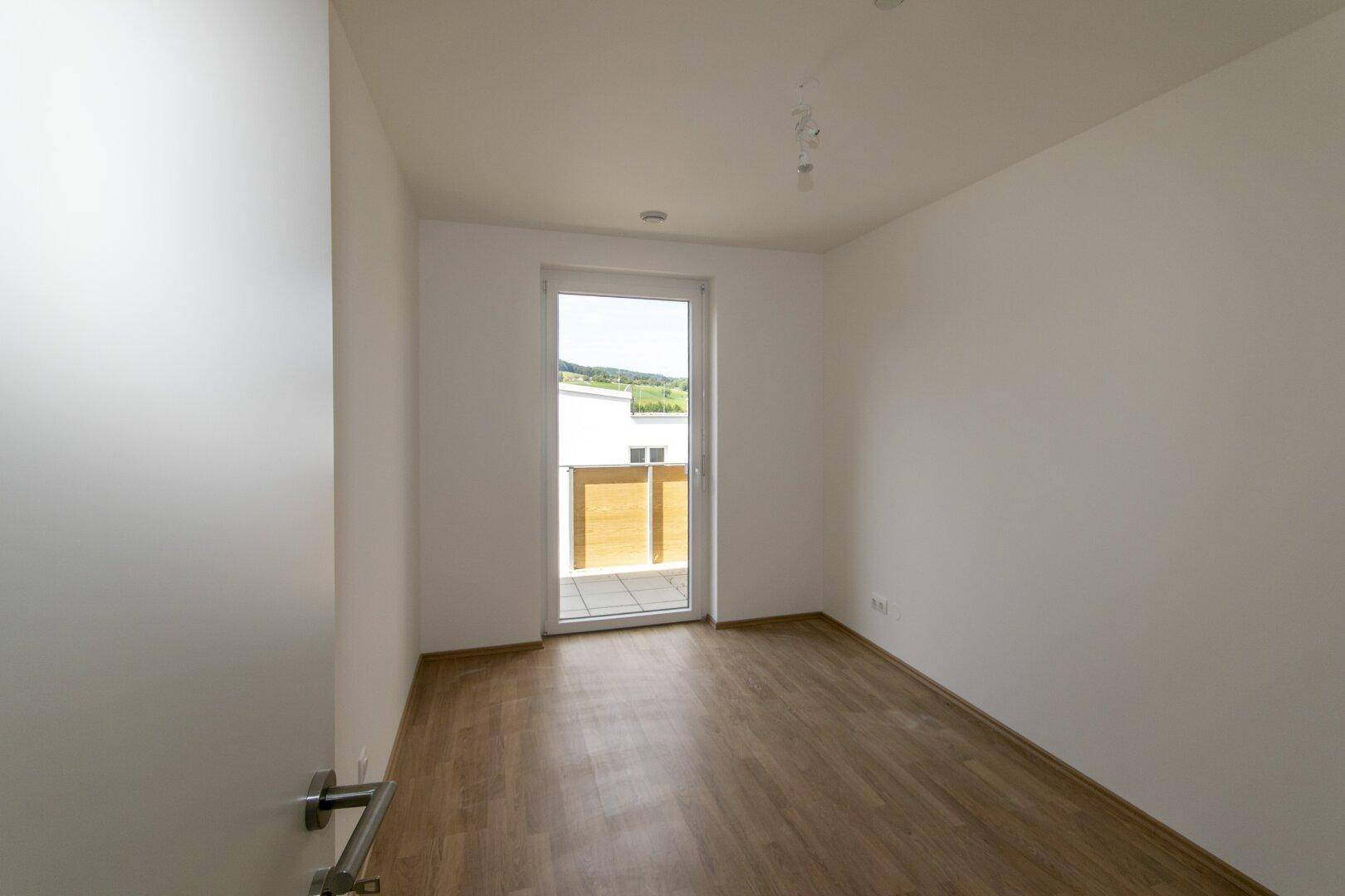 Leeres Schlafzimmer mit Laminatboden und einer Fenstertür, die auf den Balkon führt.