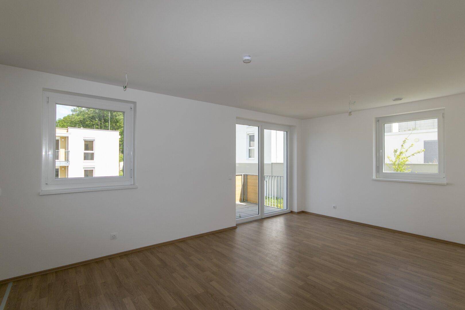 Lichtdurchfluteter Raum mit großen Fenstern und hellem Laminatboden, ideal für eine moderne Einrichtung.