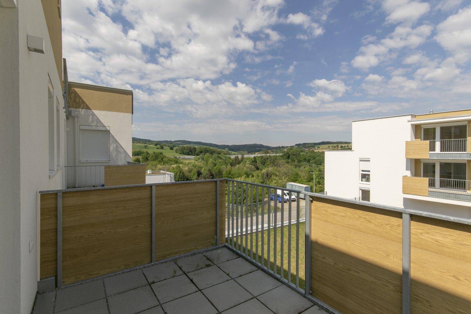 Heller Balkon mit Holzverkleidung und weitem Blick über die grüne Hügellandschaft unter blauem Himmel.