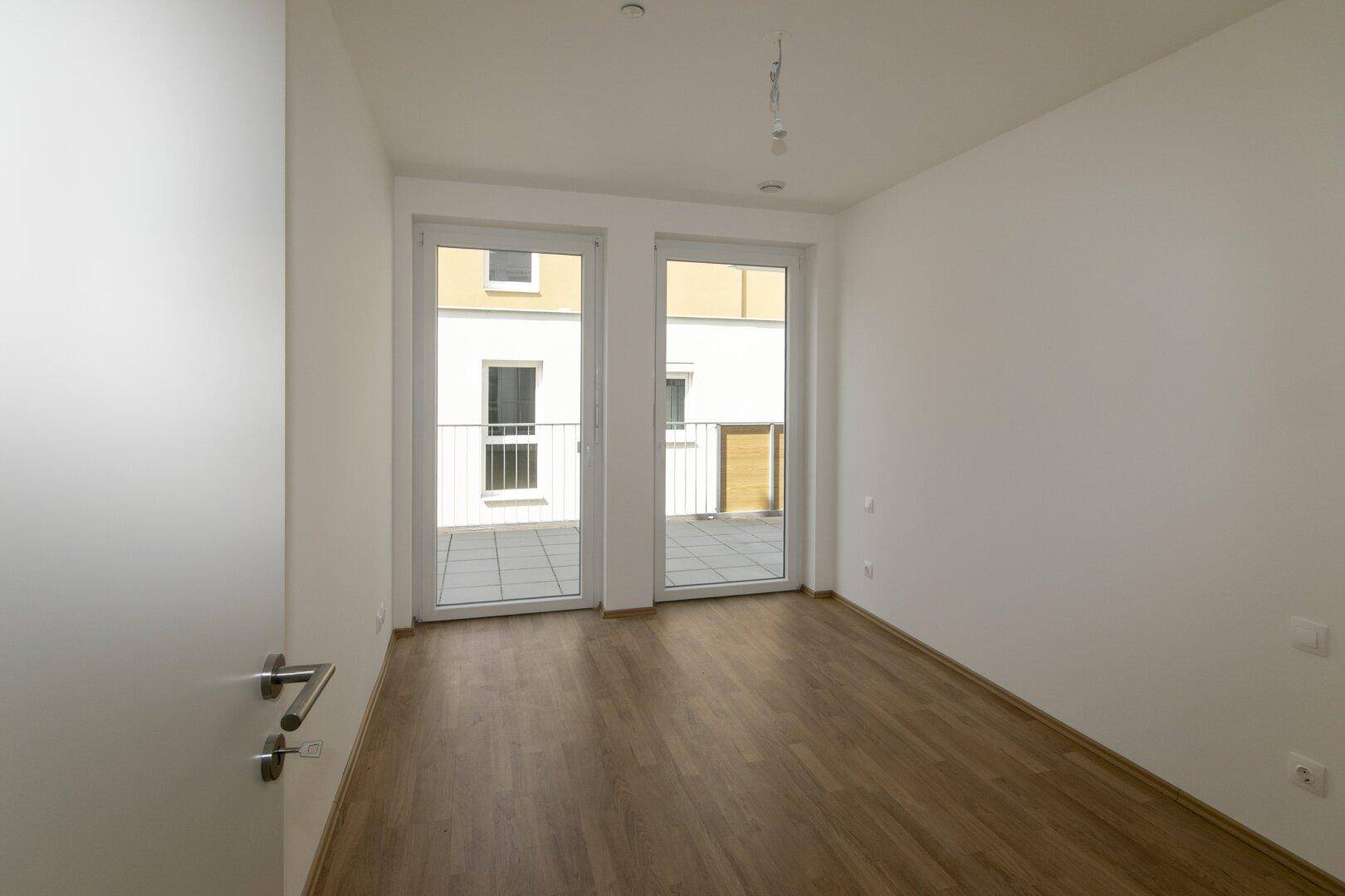 Geräumiges, helles Schlafzimmer mit Laminatboden und großen Fenstern, die auf einen Balkon führen.