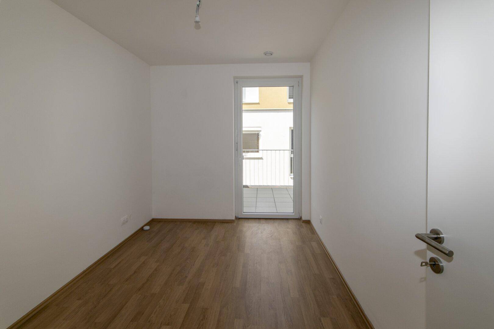 Helles Schlafzimmer mit Laminatboden und Zugang zu einem Balkon durch eine Glastür.
