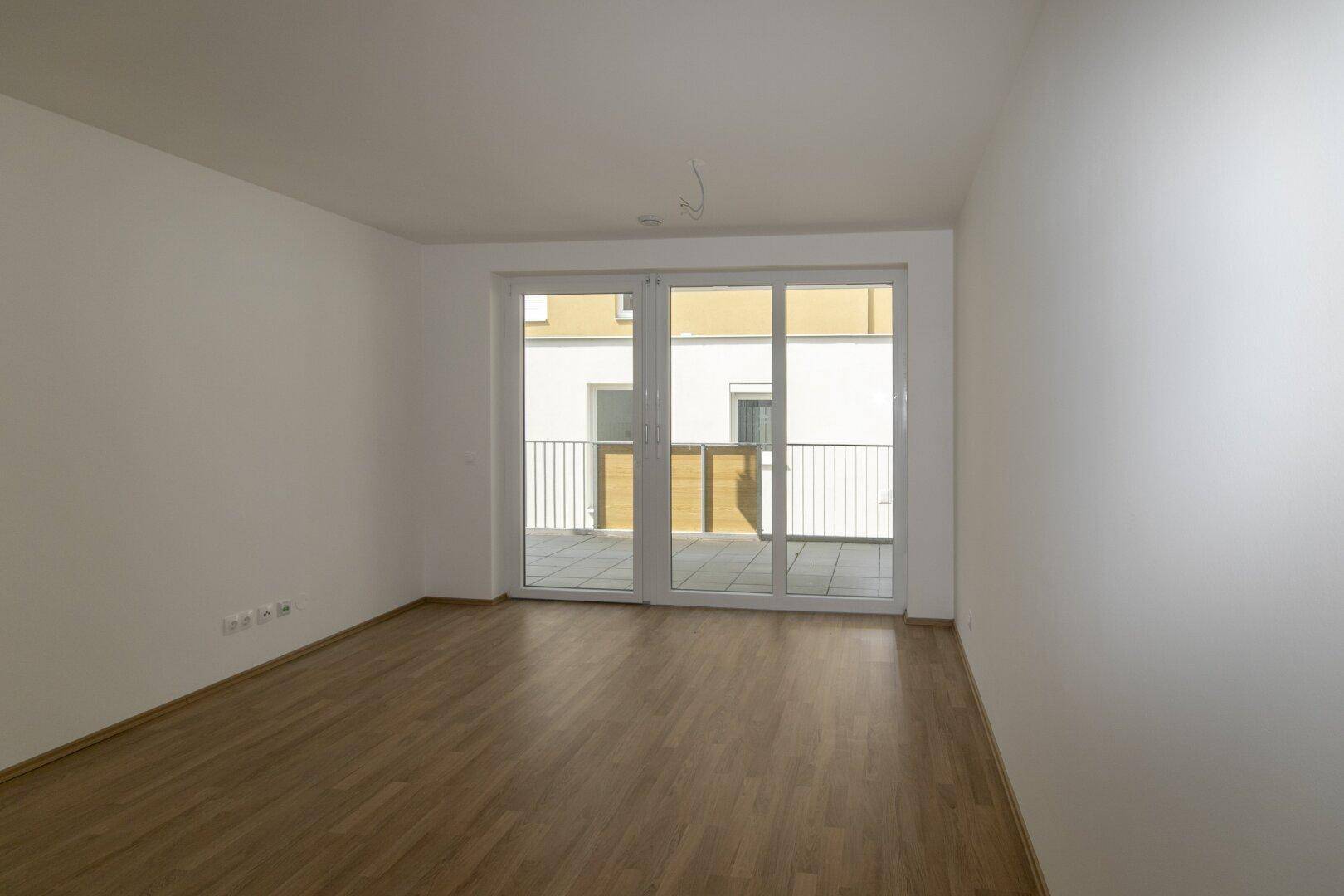 Geräumiger, heller Raum mit Laminatboden und großen Fenstern, die auf eine Terrasse führen.