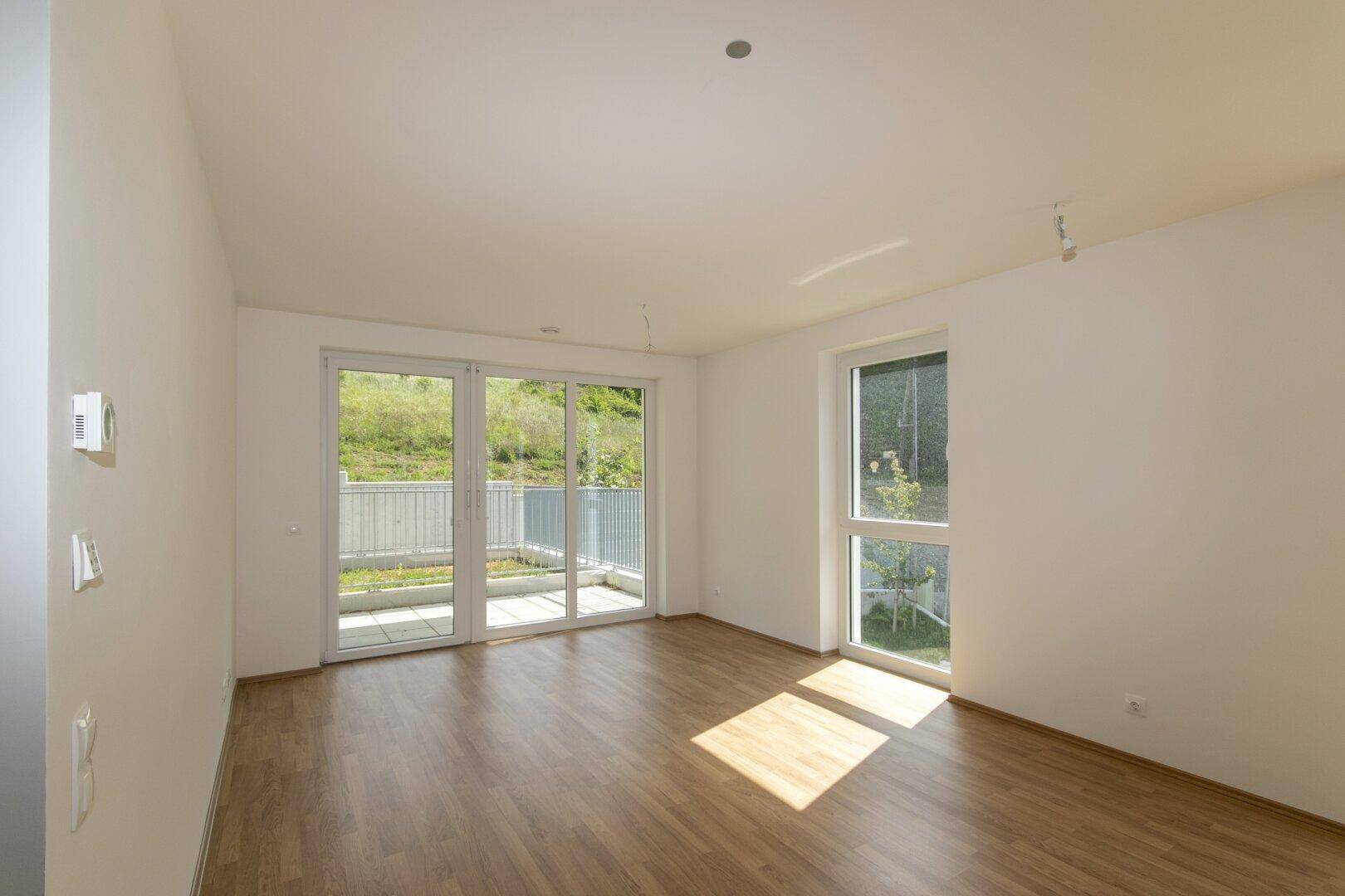 Lichtdurchflutetes Wohnzimmer mit hellem Laminatboden und großen Fenstern mit Blick ins Grüne.
