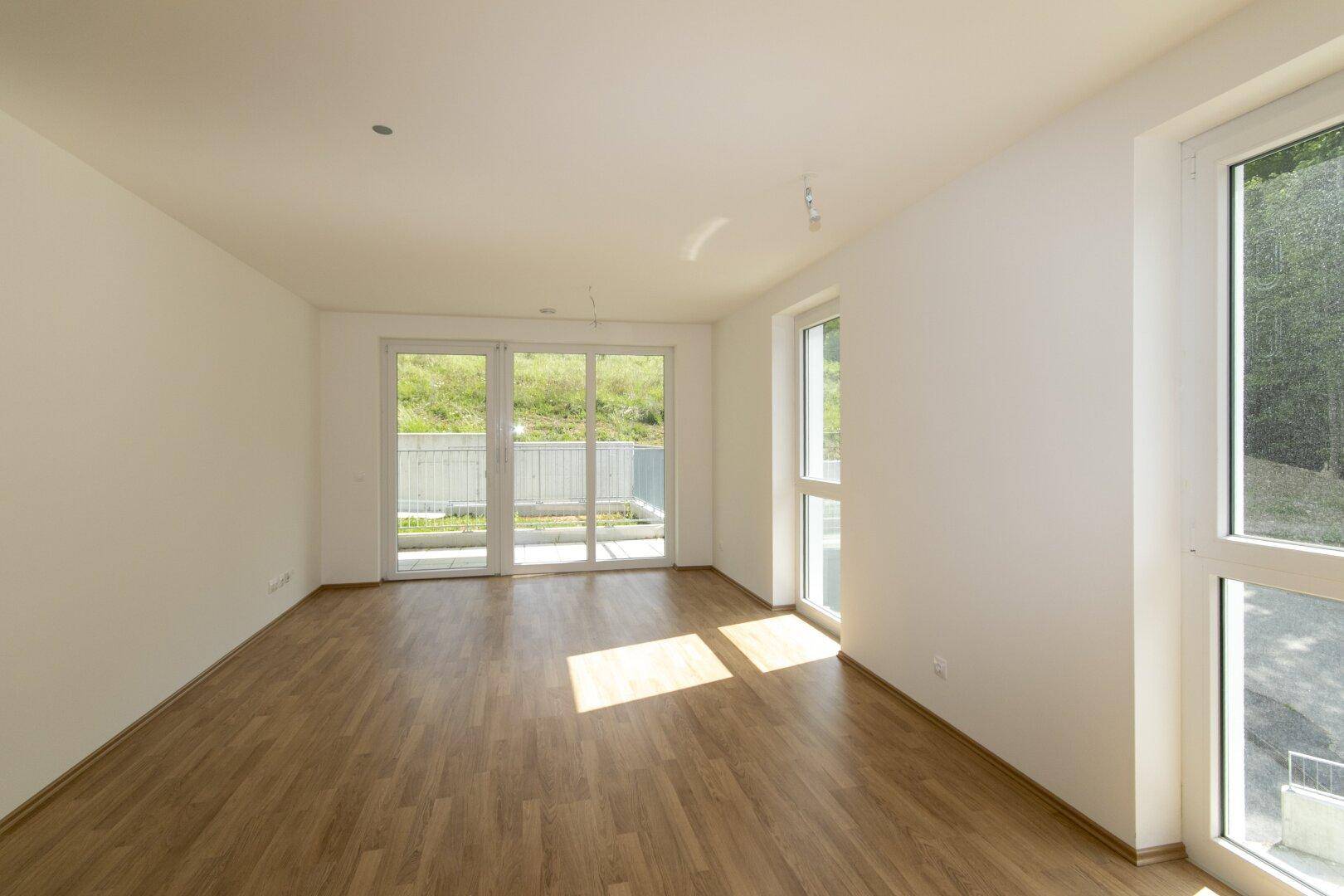 Helles, leeres Wohnzimmer mit Laminatboden, weißen Wänden und Zugang zur Terrasse.