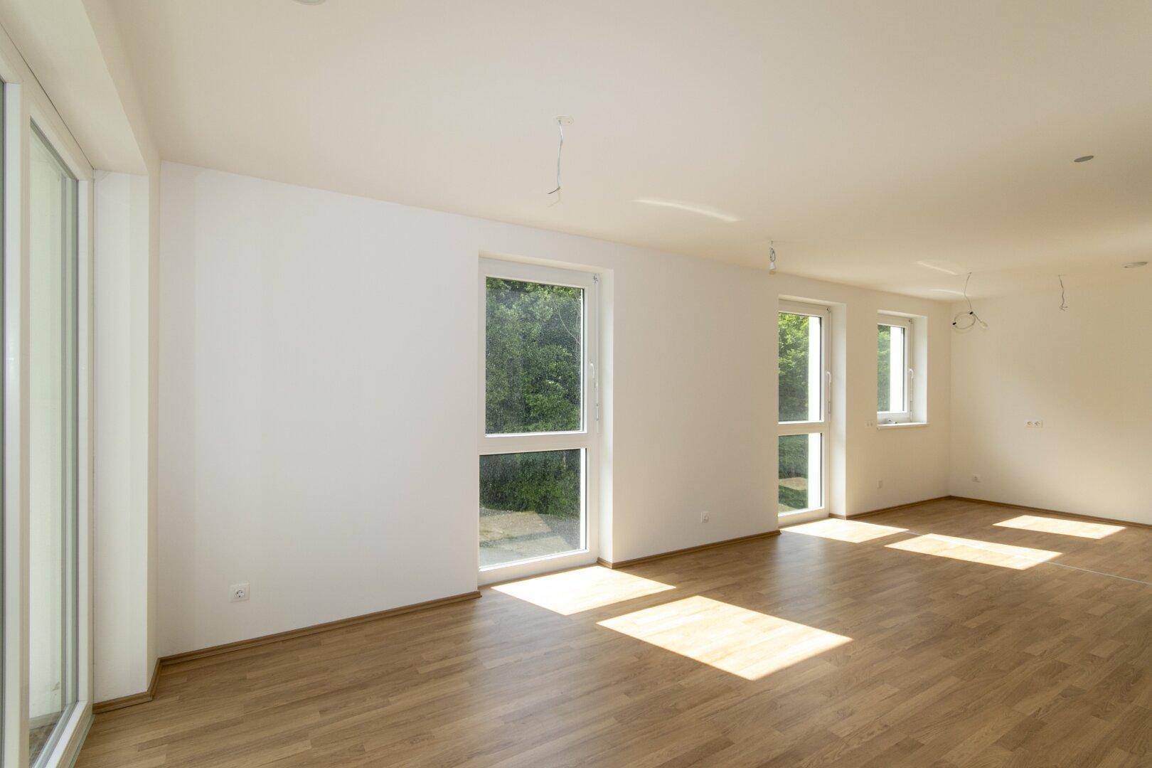 Geräumiges Wohnzimmer mit vielen Fenstern, die viel Tageslicht hereinlassen und einen grünen Ausblick bieten.
