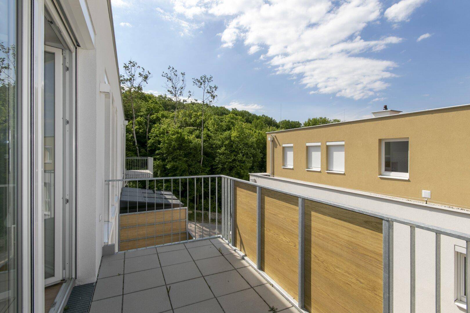Balkon mit Fliesenboden, Holzdetails und grüner Aussicht auf Bäume und Gebäude.