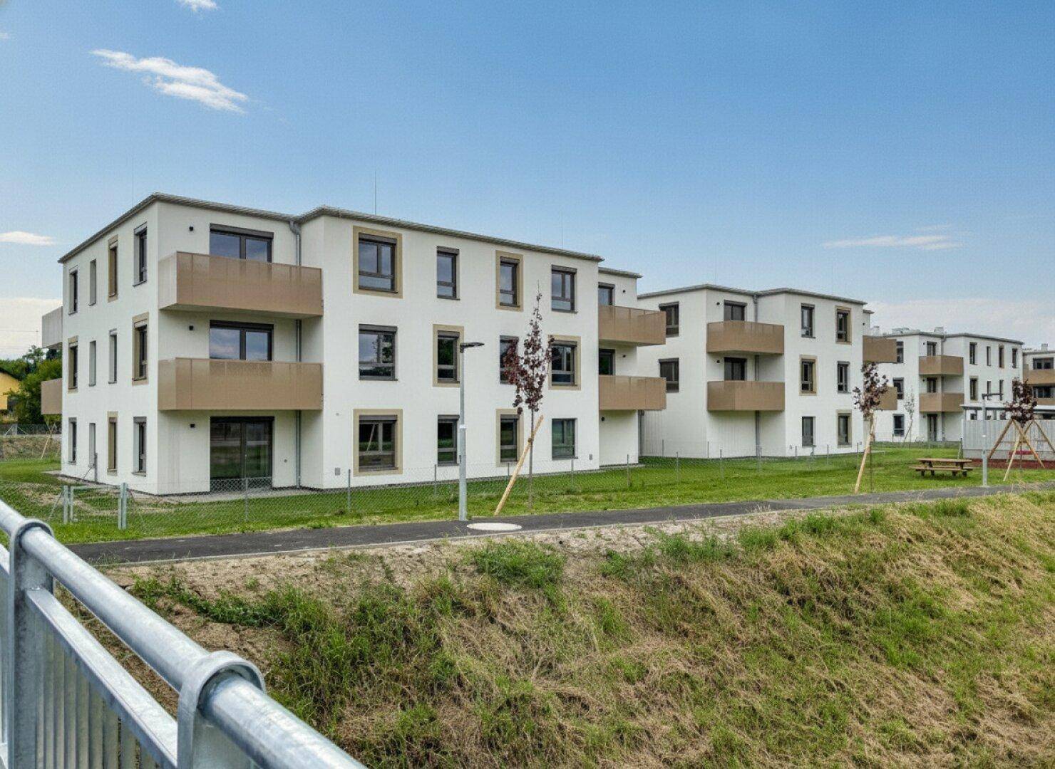 Moderne Mehrfamilienhäuser mit Balkonen und Grünflächen, umgeben von einer ländlichen Umgebung mit Spielplatz.