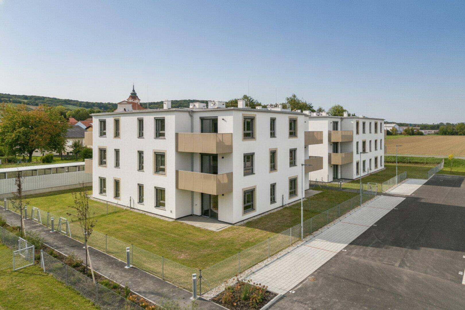 Mehrere moderne Wohngebäude mit Balkonen und gepflegten Außenanlagen, umgeben von Feldern und ländlicher Architektur.