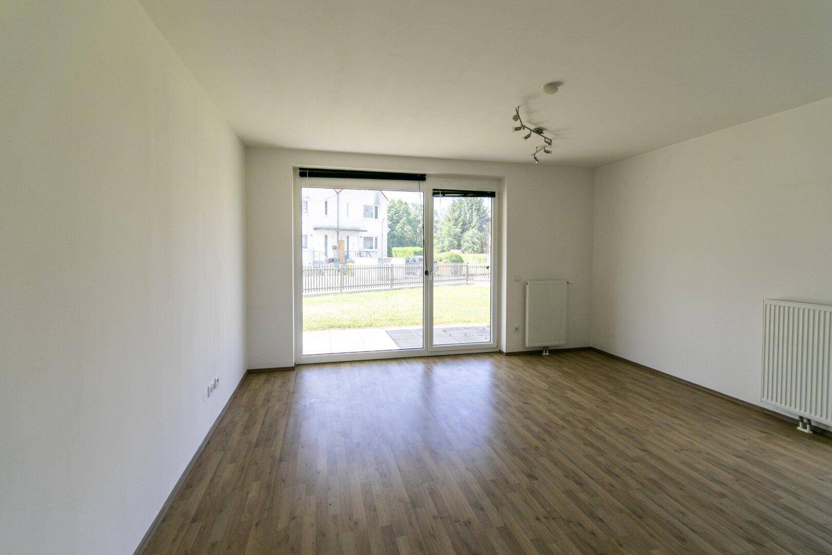 Helles, leeres Wohnzimmer mit Laminatboden, weißen Wänden und Zugang zur Terrasse.