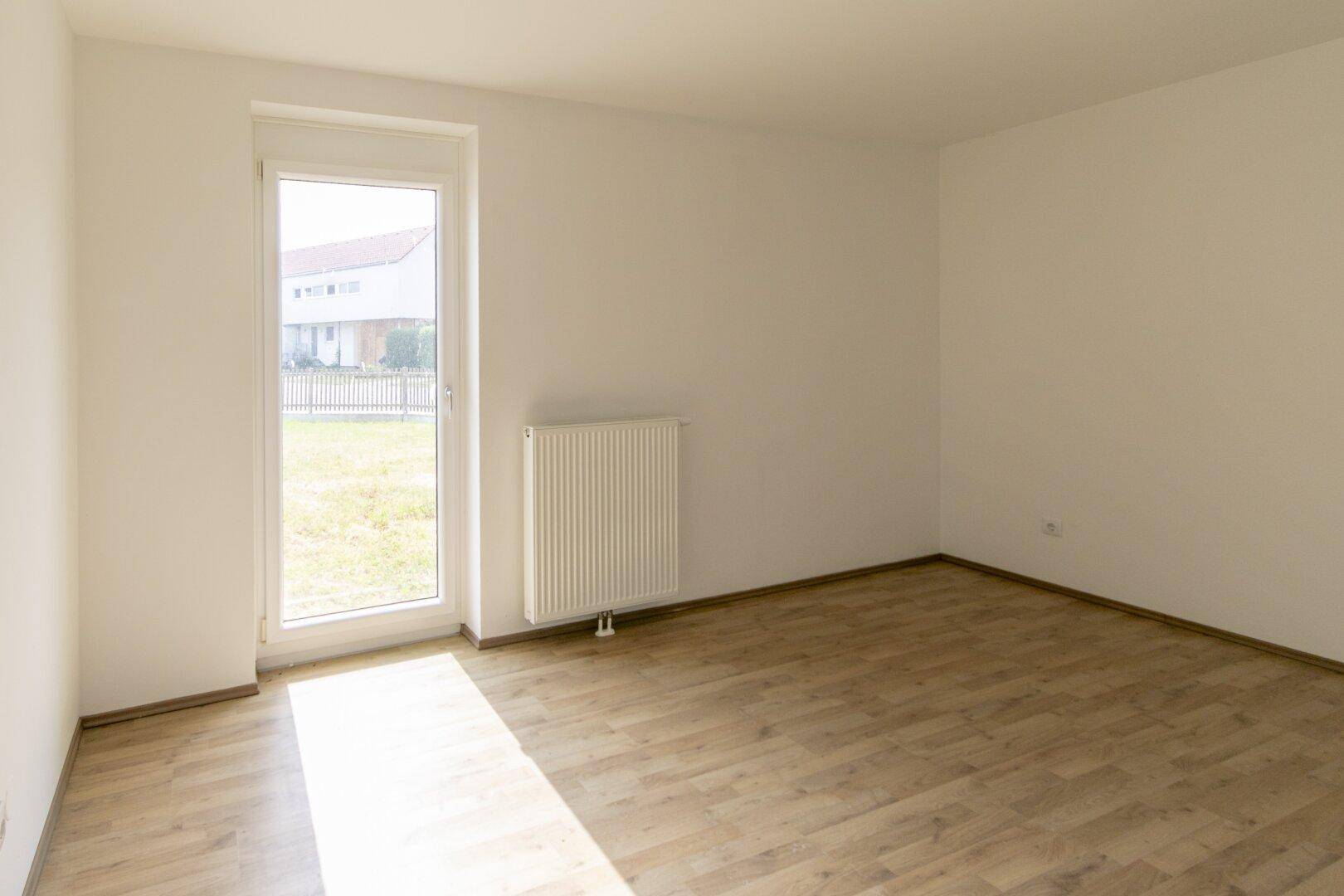 Sonniger, leerer Raum mit Laminatboden, weißen Wänden und einem Fenster mit Blick ins Grüne.