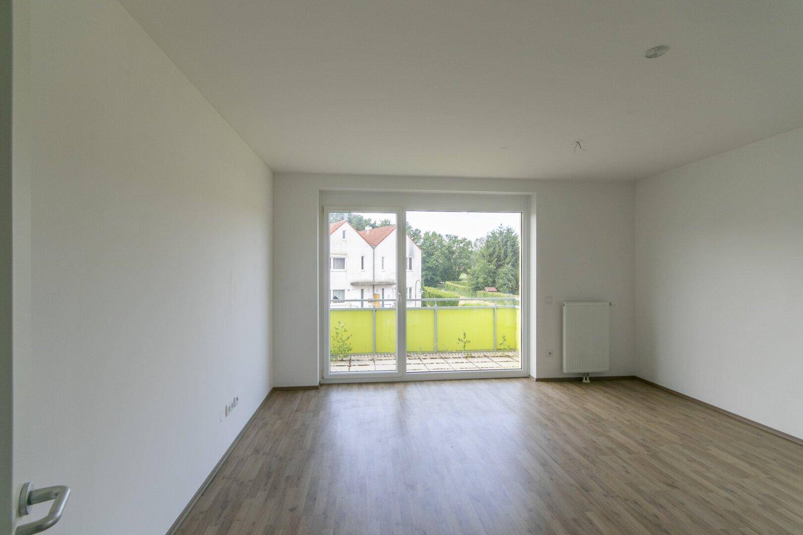 Heller Wohnraum mit Laminatboden, weißen Wänden und großem Fenster mit Zugang zum Außenbereich.