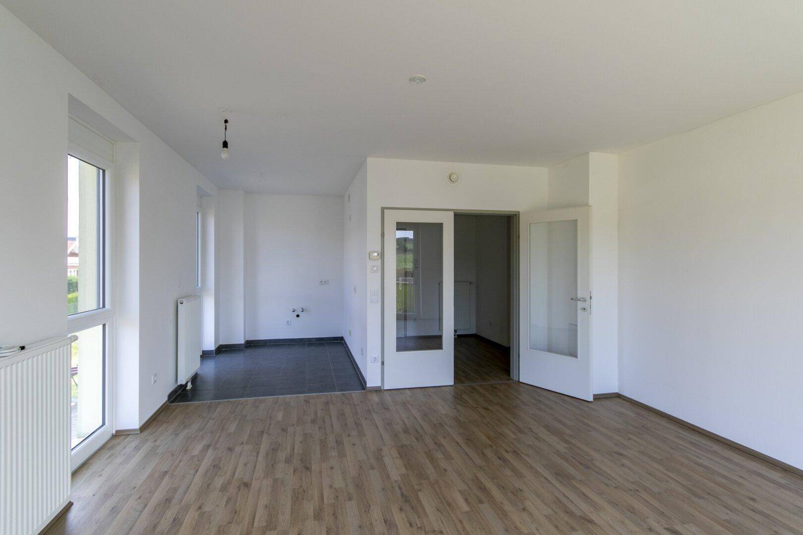 Lichtdurchfluteter Wohnraum mit Holzlaminat und offenem Übergang zu einem weiteren Zimmer.