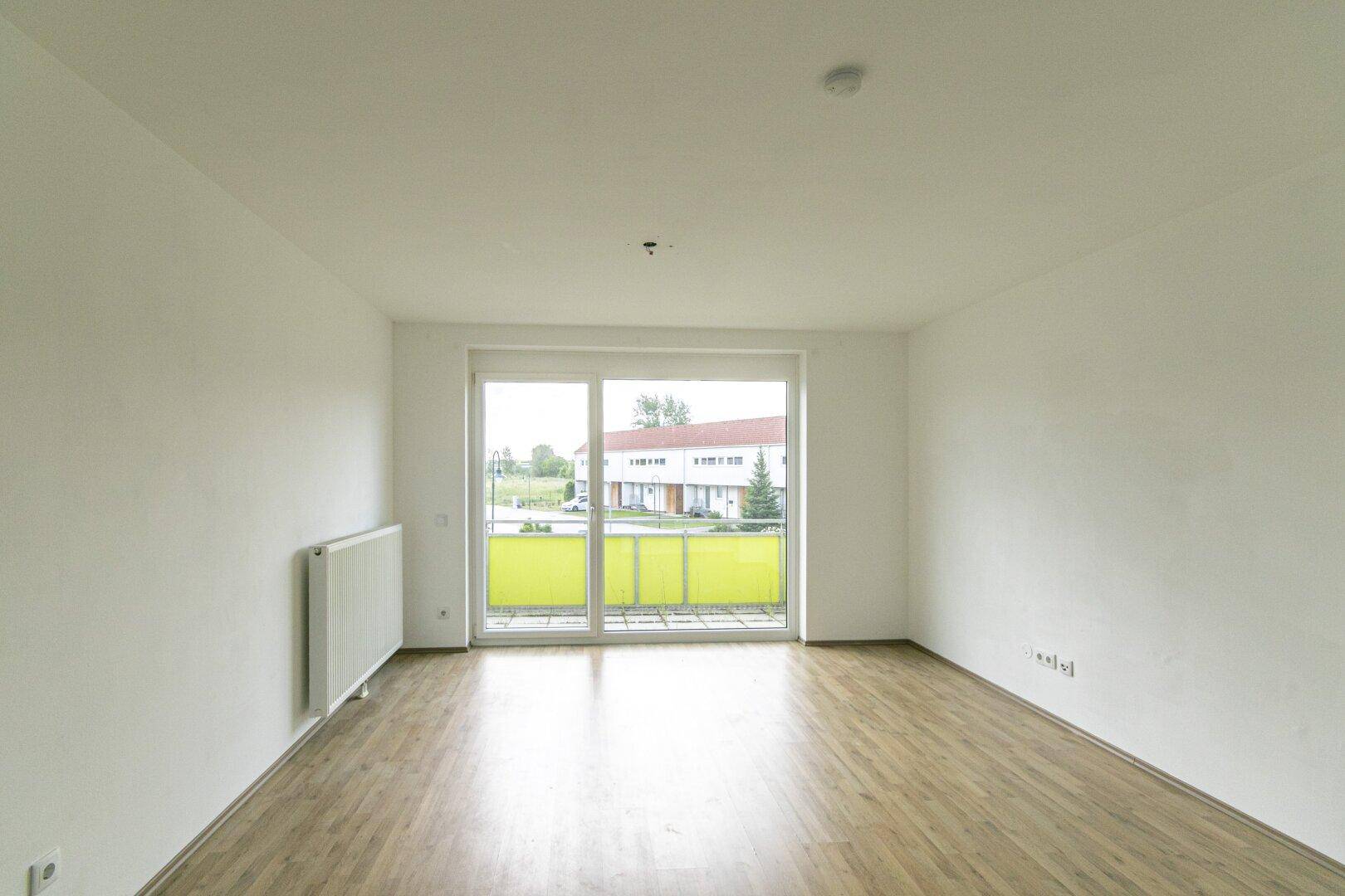 Leeres Wohnzimmer mit Laminatboden und großer Balkontür, die viel Tageslicht hereinlässt.