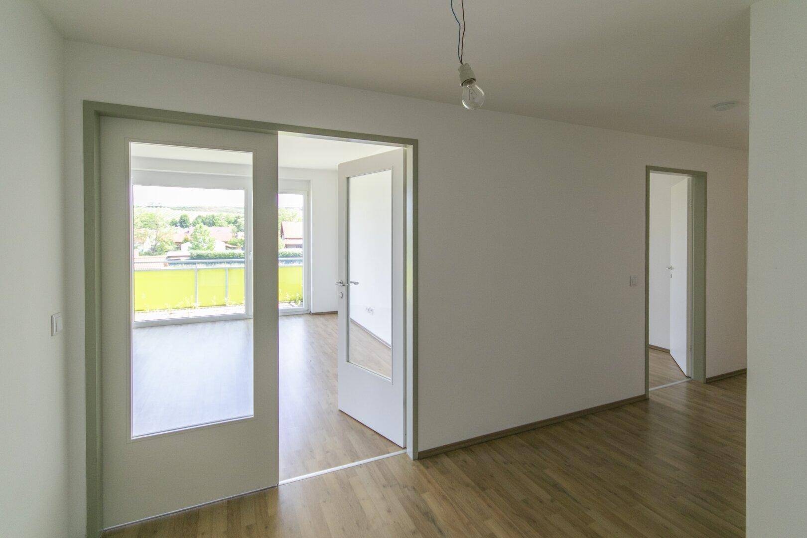 Heller Raum mit Laminatboden und Zugang zu einem weiteren Zimmer mit Balkonblick.