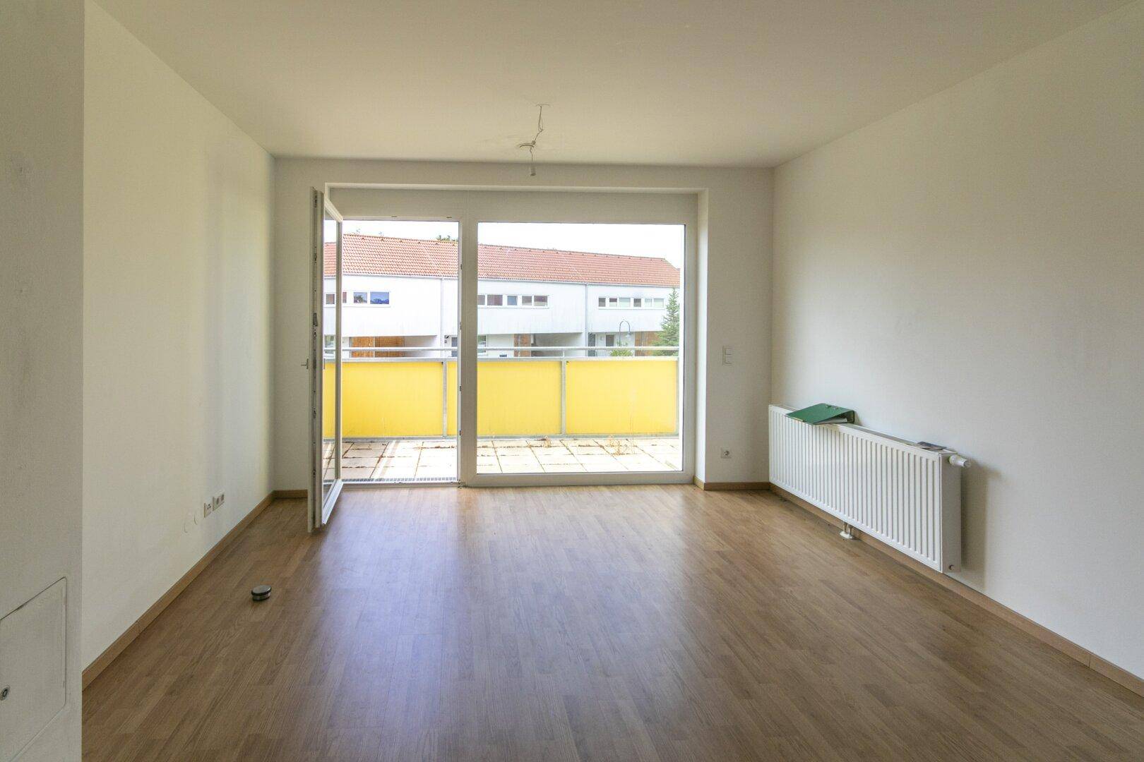 Helles, leeres Wohnzimmer mit Laminatboden und Zugang zu einem Balkon.