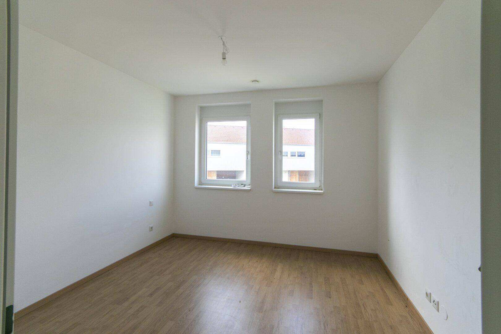 Leeres Schlafzimmer mit zwei Fenstern, Laminatboden und hellen Wänden.