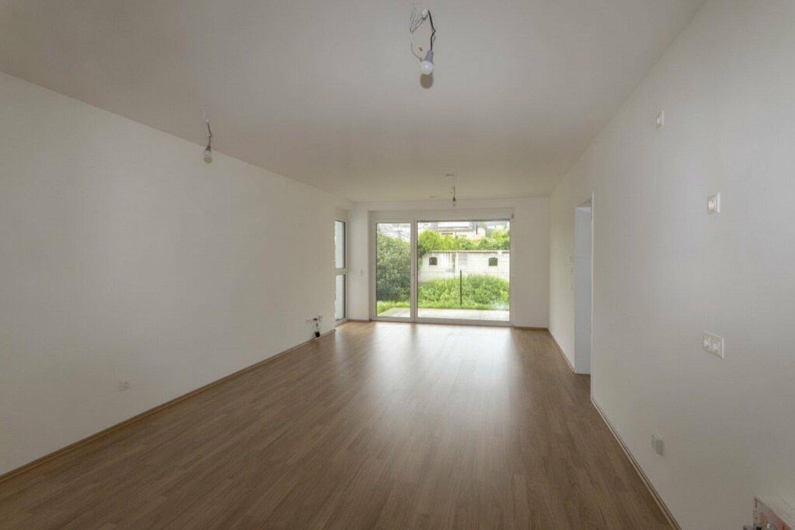 Helles, leeres Wohnzimmer mit Holzboden und großer Schiebetür zum Außenbereich.