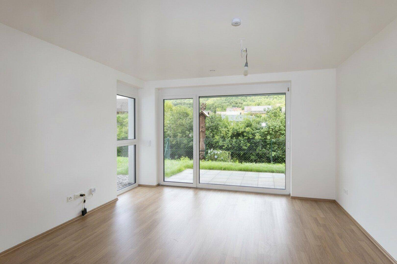Helles, leeres Wohnzimmer mit Holzboden und großem Fenster mit Zugang zur Terrasse.