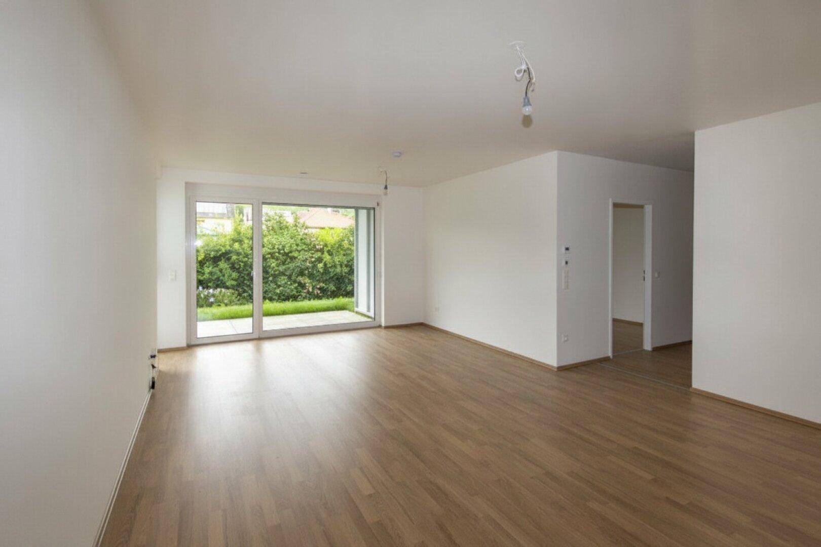 Geräumiges, helles Wohnzimmer mit Laminatboden und Blick ins Grüne durch große Fenster.