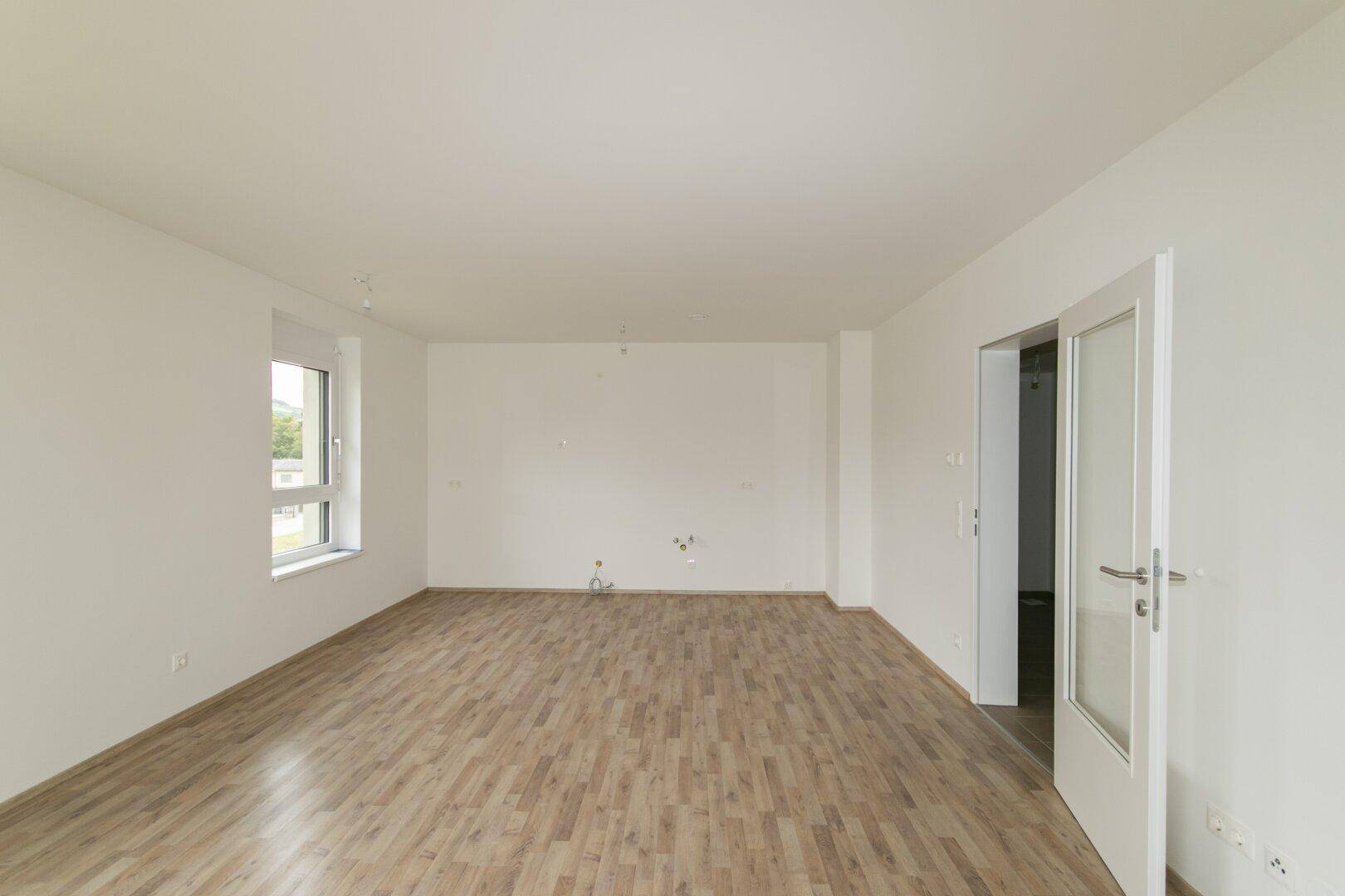 Leeres Wohnzimmer mit Laminatboden, weißen Wänden und viel Tageslicht durch die Fenster.