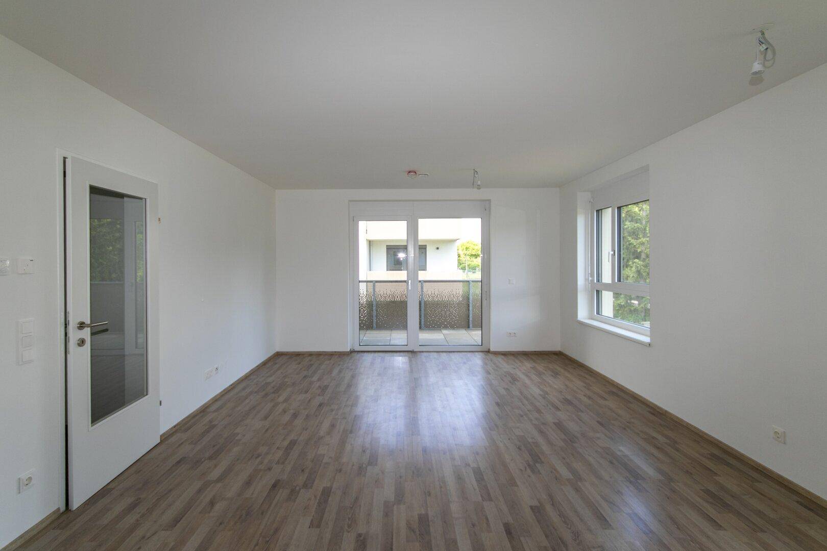 Geräumiges, leeres Wohnzimmer mit Laminatboden, weißen Wänden und Zugang zum Balkon.