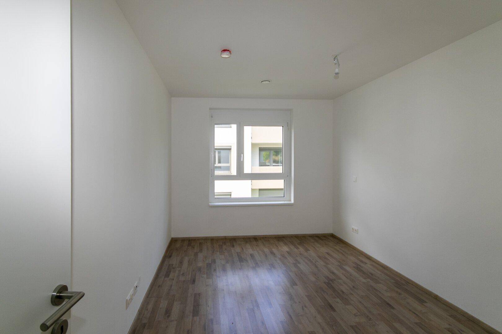 Helles Schlafzimmer mit Laminatboden und Blick aus dem Fenster auf die Umgebung.