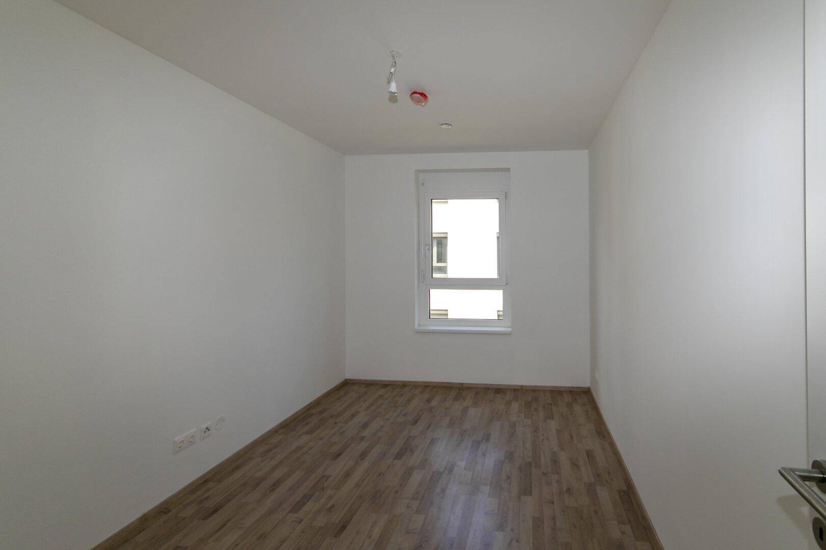 Leeres Schlafzimmer mit Laminatboden und einem Fenster, das viel Tageslicht hereinlässt.