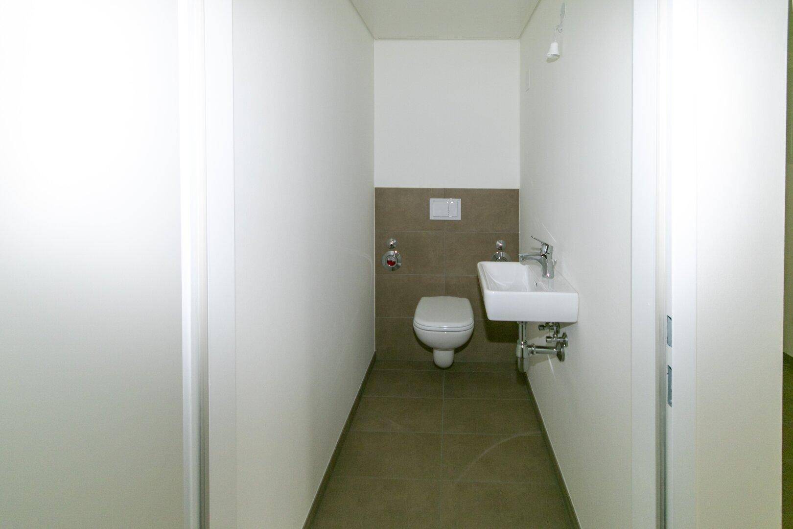 Modernes Gäste-WC mit wandhängender Toilette, kleinem Waschbecken und neutralen Fliesen.