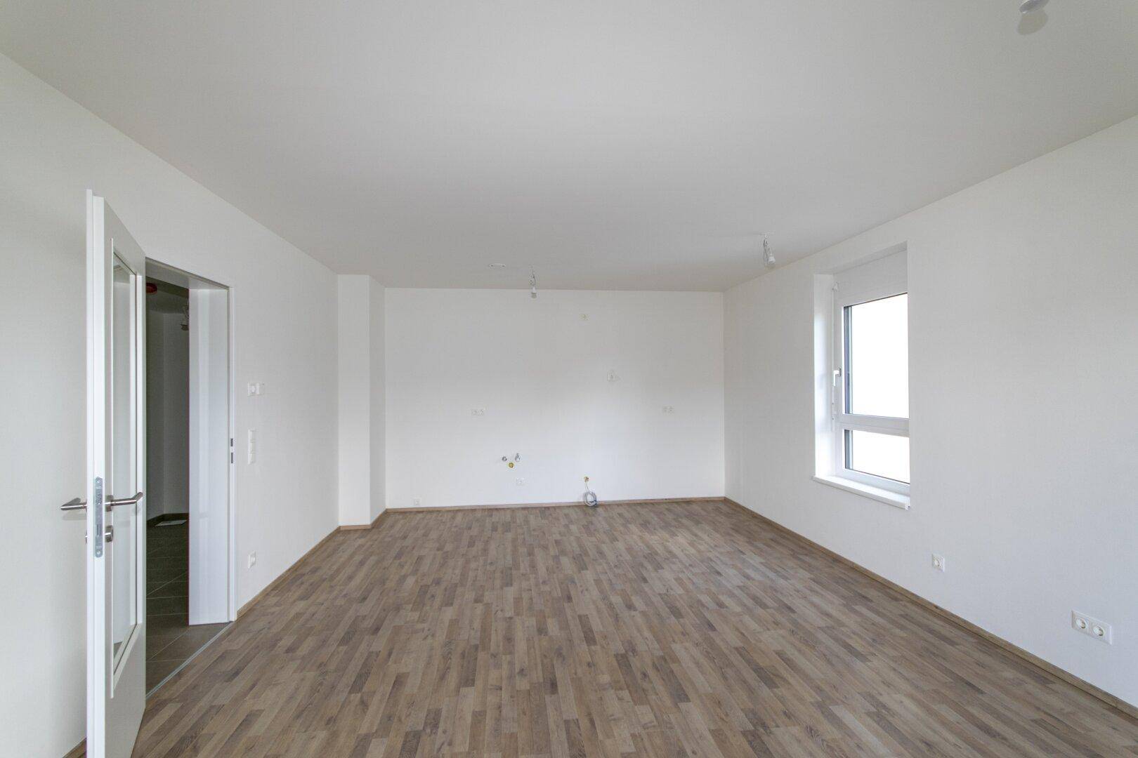 Leeres Wohnzimmer mit Laminatboden, weißen Wänden und einem Fenster, bereit zur individuellen Gestaltung.