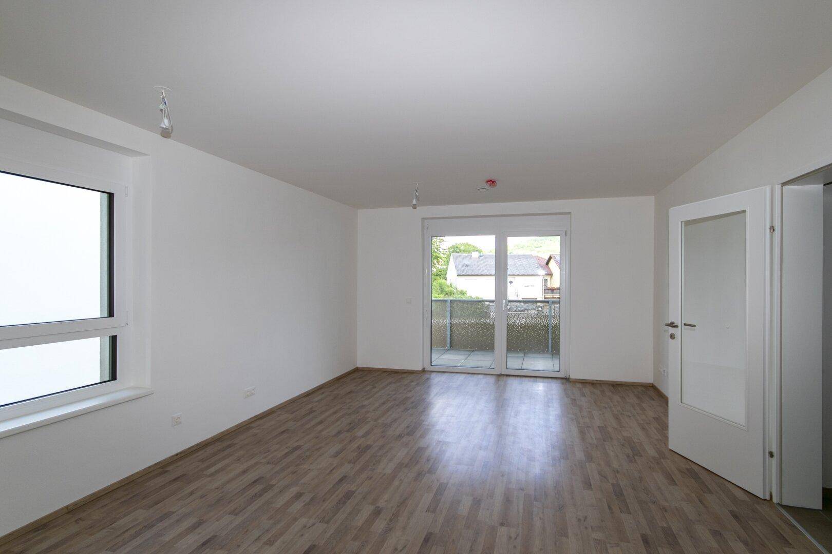 Helles, leeres Wohnzimmer mit Laminatboden, weißen Wänden und Zugang zum Balkon.