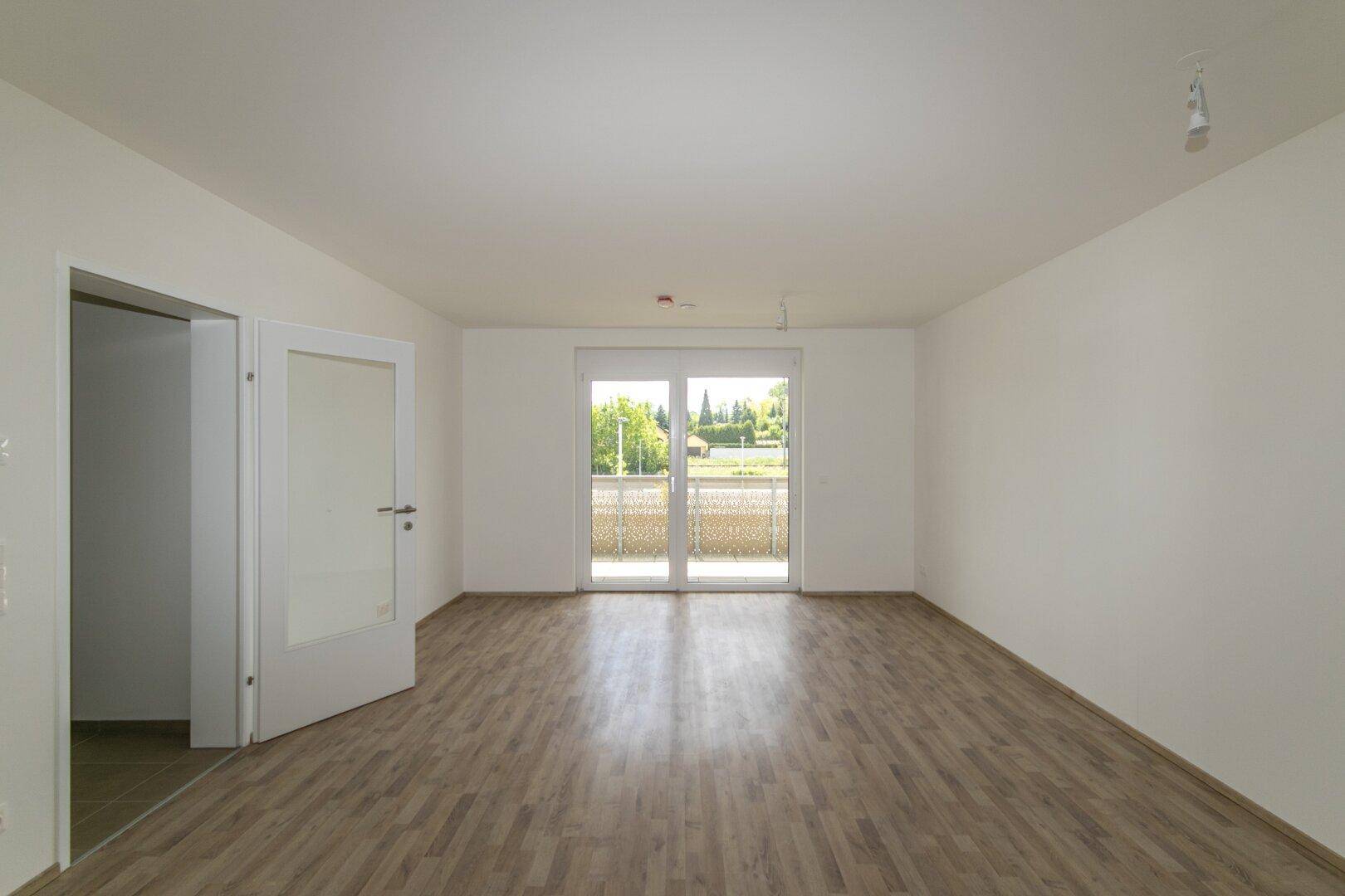 Helles, leeres Zimmer mit Laminatboden und Zugang zu einem Balkon.