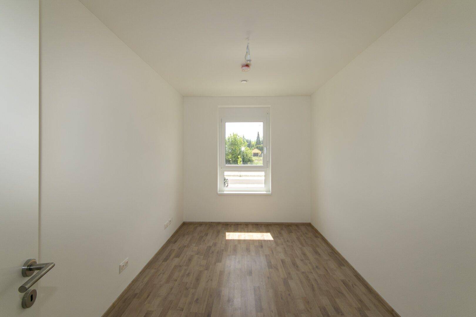 Helles, leeres Zimmer mit Laminatboden und einem Fenster mit Blick ins Grüne.