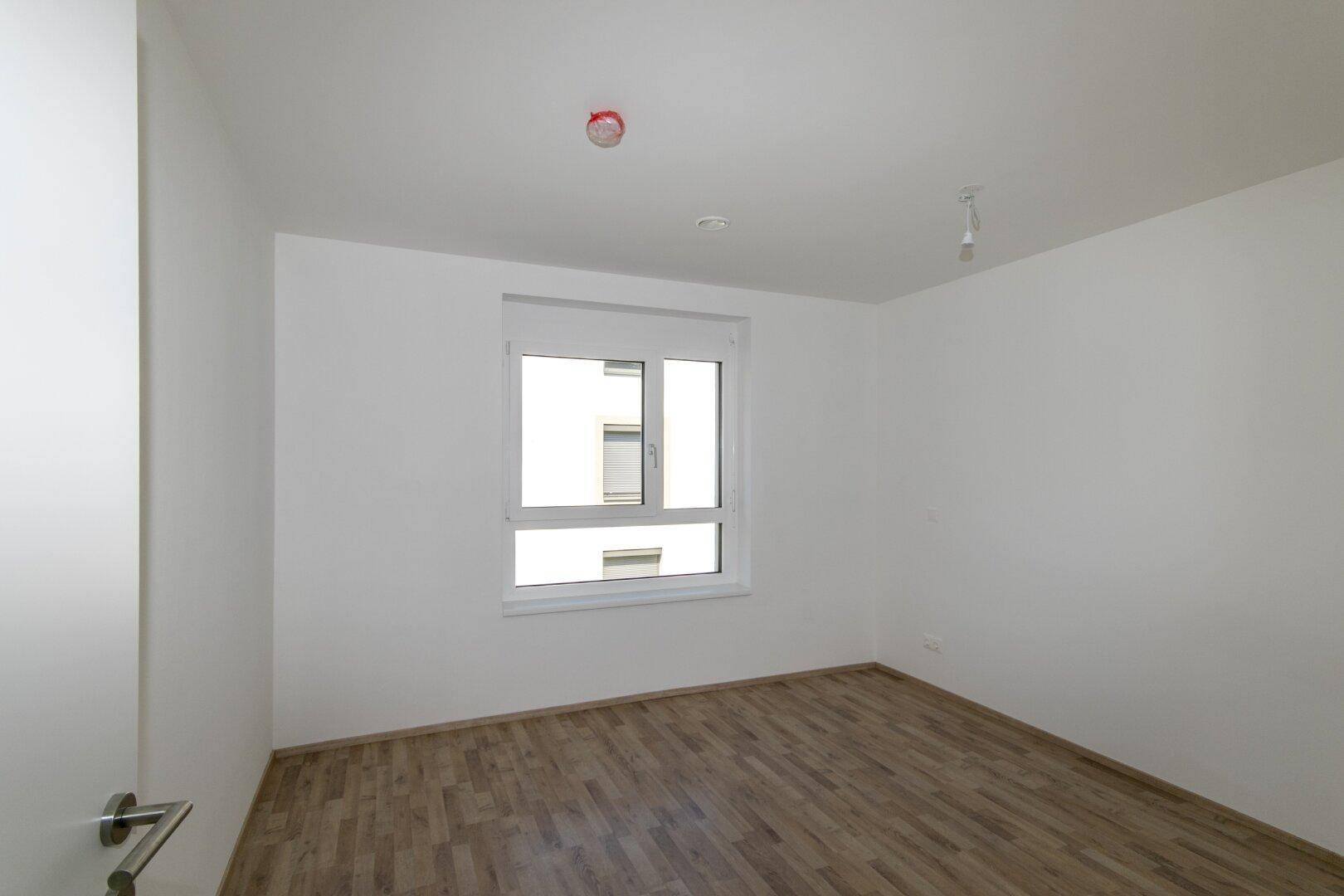 Helles Schlafzimmer mit Laminatboden und einem Fenster, das viel Tageslicht hereinlässt.