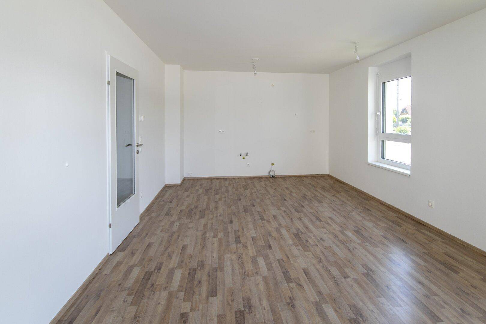 Leerer Küchenraum mit Laminatboden, weißen Wänden und vorbereiteten Anschlüssen.