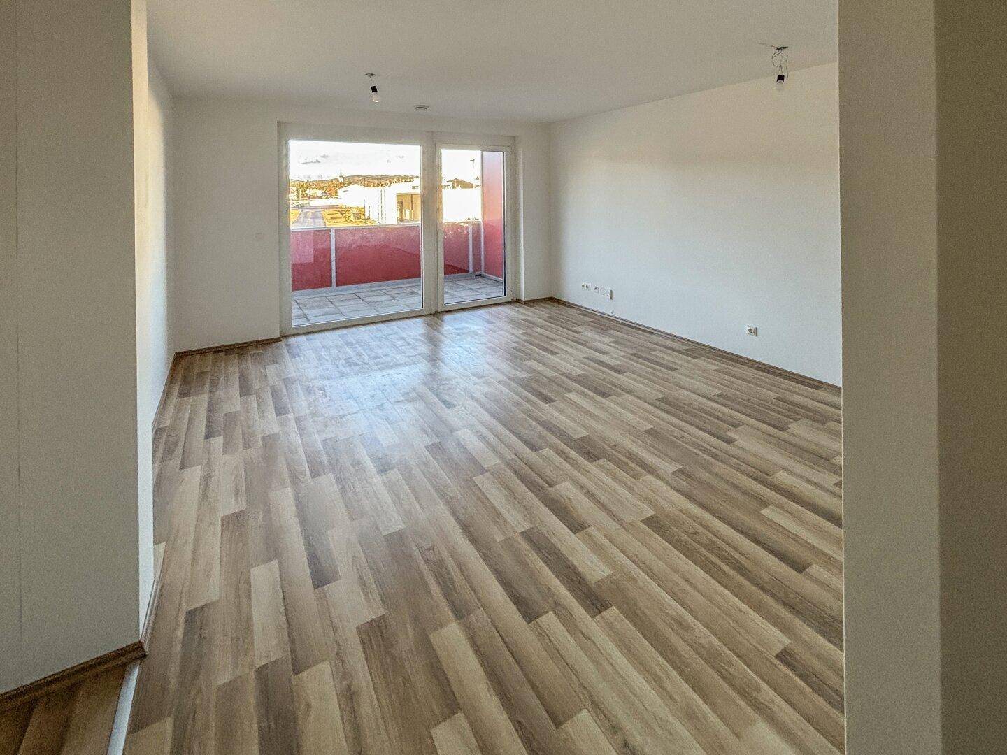 Helles, leeres Wohnzimmer mit Laminatboden und Zugang zu einem Balkon.