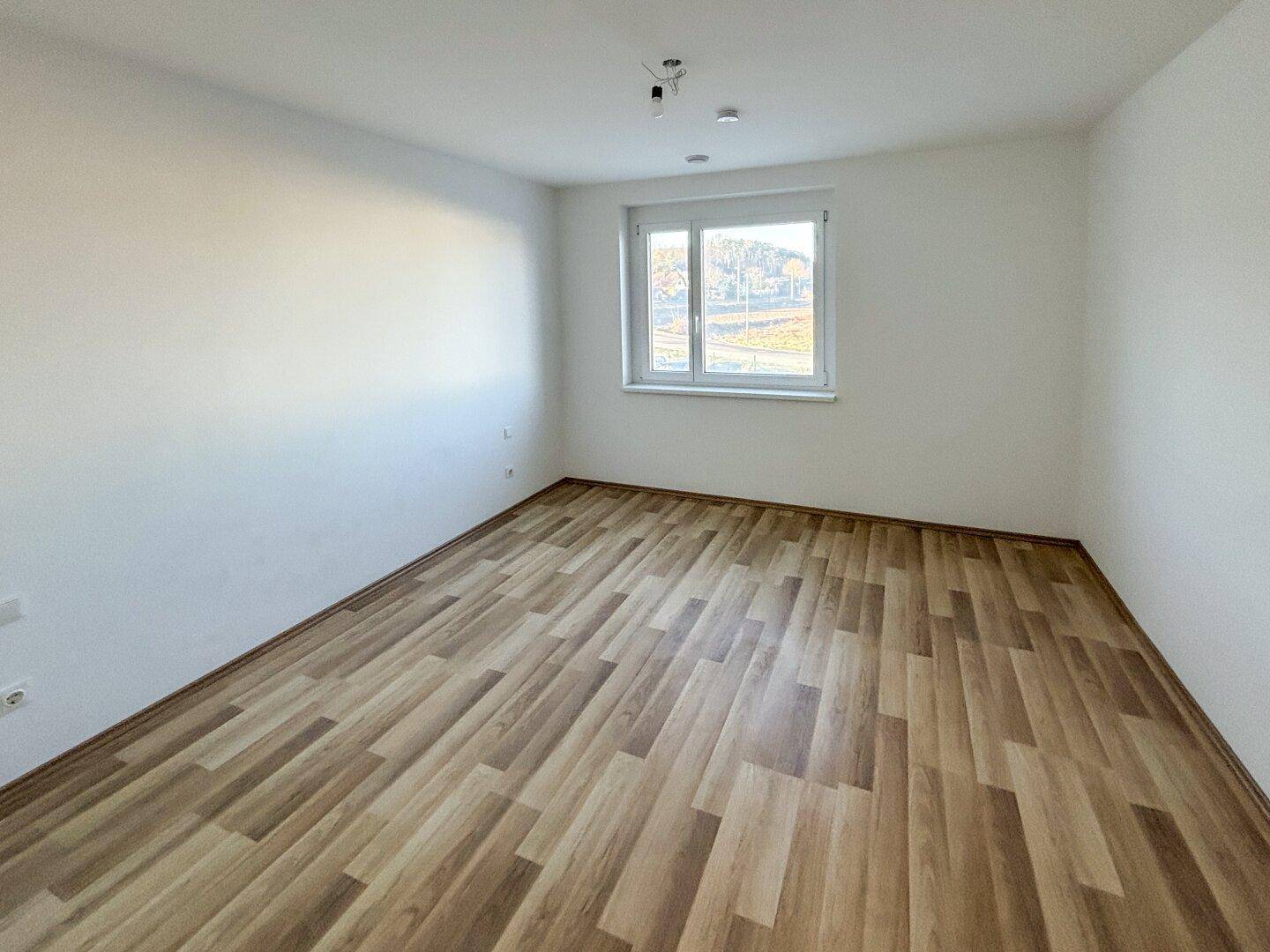 Helles Schlafzimmer mit Laminatboden und einem Fenster mit Blick ins Grüne.