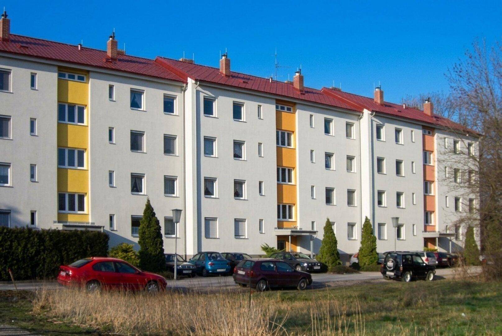 Lange Reihe von Mehrfamilienhäusern mit weißer Fassade, roten Dächern und Parkplätzen davor.