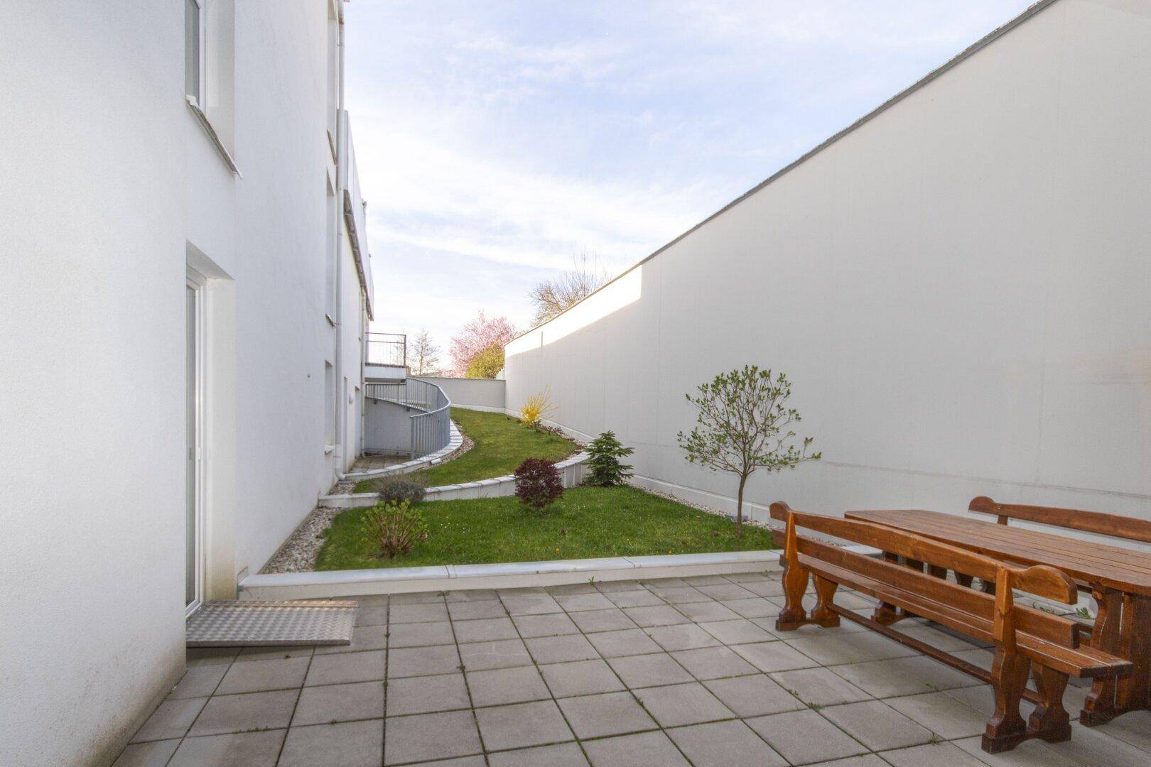 Kleiner Gartenbereich mit Terrasse und Sitzgelegenheiten, umgeben von weißen Wänden und Grünflächen.