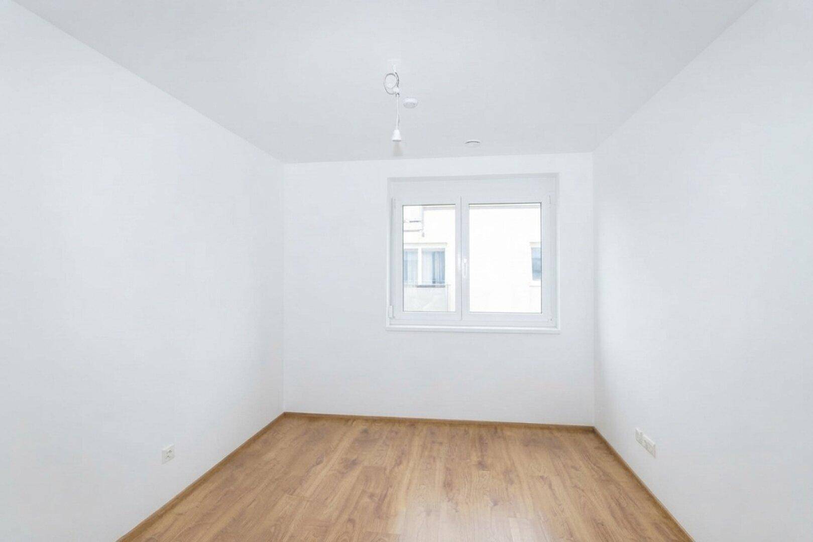 Leeres Schlafzimmer mit Laminatboden und einem großen Fenster, das viel Tageslicht bietet.