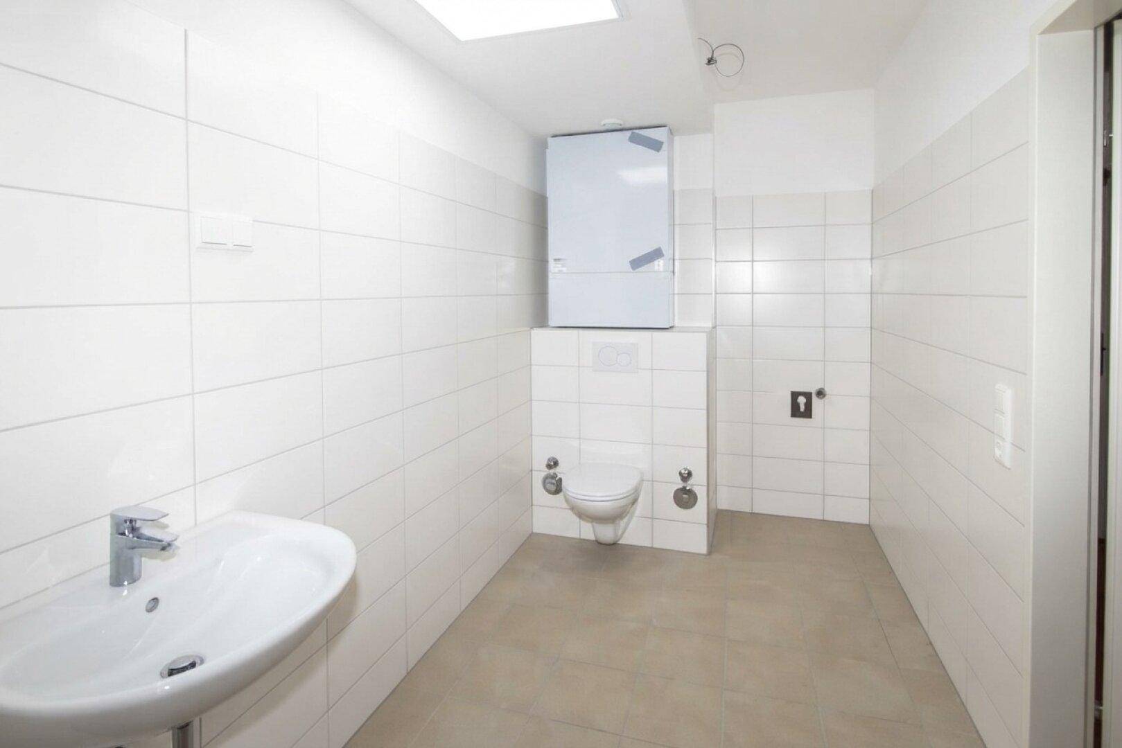 Helles Badezimmer mit Wand-WC, Waschbecken und Boiler, komplett mit weißen Fliesen ausgestattet.
