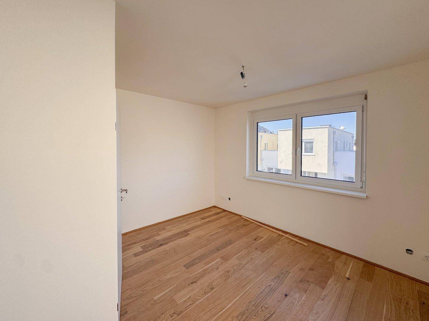 Helles Schlafzimmer mit Holzboden und Fenster, das einen Blick auf die Nachbargebäude bietet.