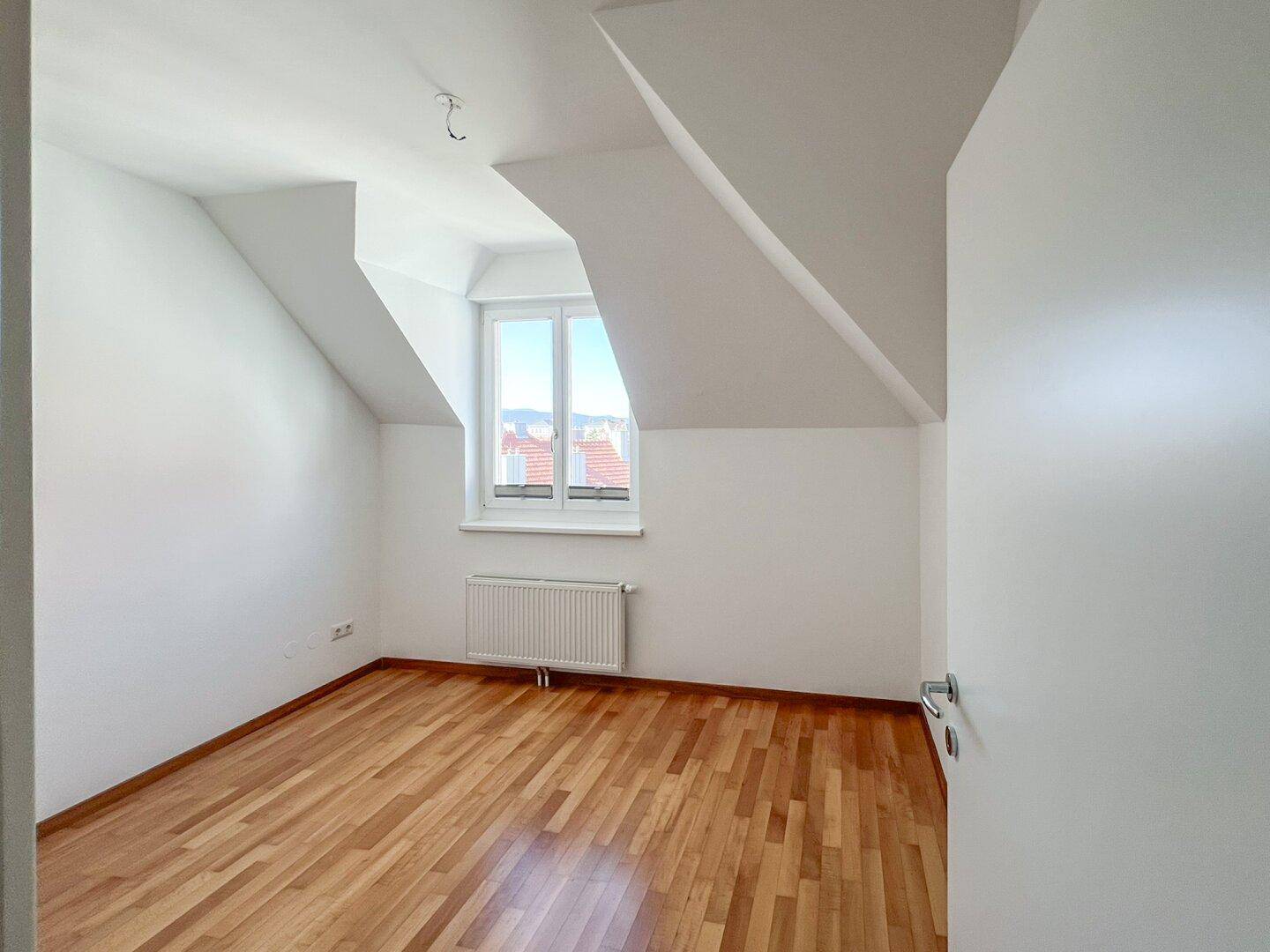 Dachgeschosszimmer mit Holzboden, schrägen Wänden und einem Fenster, das viel Licht hereinlässt.